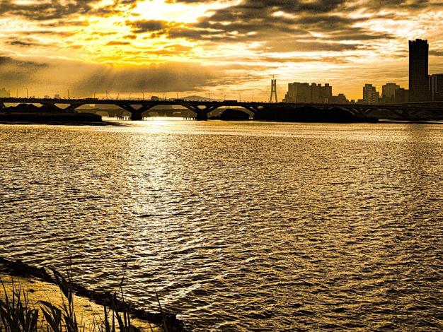 傍晚時分，夕陽緩緩自橋邊落下，淡水河面灑染著金黃色彩